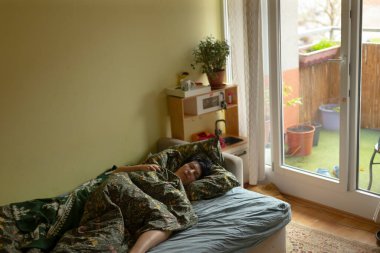 Genç adam balkonun yanındaki yatakta uyuyor, kamuflaj ceketi giyiyor, güneş ışığı sürgülü kapıdan içeri akıyor, raftaki saksı bitkileri dağınık kutular ve fotoğraflar, yatağın yanındaki rahat botlar,