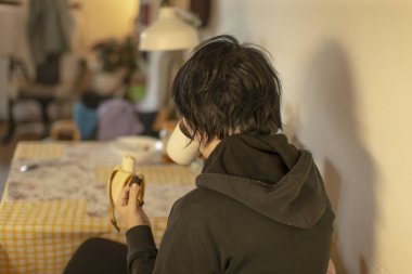 Mutfak masasında oturan, muz yiyen, koyu renk kapüşonlu kapüşonlu giyen, kısa siyah saçlı, kareli masa örtüsü, sıcak lamba, masa örtüsü ve tabak, ev yemeği, öğrenci.