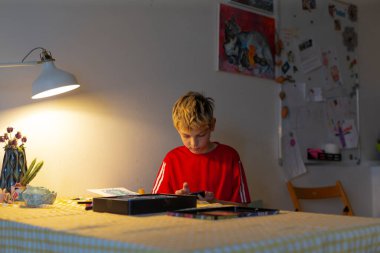 Beyaz çocuk mutfak masasına odaklanmış ev ödevlerini lambanın ışıltısı, kırmızı tişört, açık defter ve kalem, rahat iç mekan, masada fincan ve atıştırmalıklar, sessiz konsantrasyon altında yazıyor.