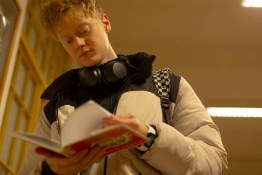 Beyaz öğrenci sıcak ışıklar altında kitap okuyor, kulaklıklar omuzlarda dinleniyor, sayfalara özen gösteriliyor, ders kitabını açık tutuyor ve notlar alıyor, akademik atmosfer ve sessizlik.