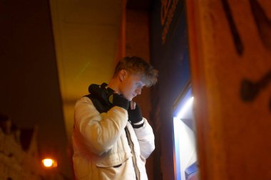 Bankamatik büfesindeki genç beyaz adam neon ışığı altında para çekiyor, beyaz kirpi ceketi ve kulaklık takıyor, eldivenli eller kart giriyor, sokak arkası, soğuk akşam atmosferi.,