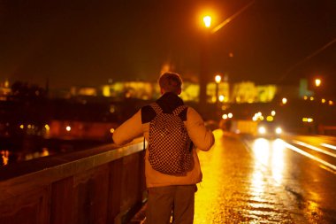Gezgin gece aydınlatılmış köprüyü seyreder, tek tek nehri geçer parıldayan şehir manzarasına doğru, Sırt çantalı kişi parlayan köprüyü ve uzak şehir ışıklarını düşünür