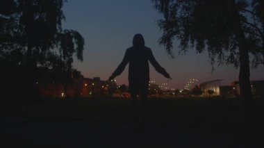 Akşam parkında şehir ışıklarına karşı antrenman yapan bir sporcu, sakin gökyüzü altında ip atlayan bir koşucu silueti, huzurlu bir akşam parkında antrenman yapmak, hareketin altını çizmek ve şehrin ambiyansı.