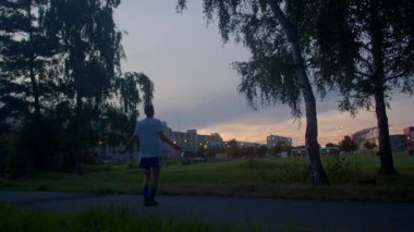 Şehir parkında alacakaranlıkta egzersiz yapan bir adam, ufuk çizgisini, sakin duruşu, uzak binaları çerçeveleyen huş ağaçlarıyla alacakaranlık atmosferini, yansıtıcı hızı takdir etmek için yol kenarında hafif koşular yapıyor.