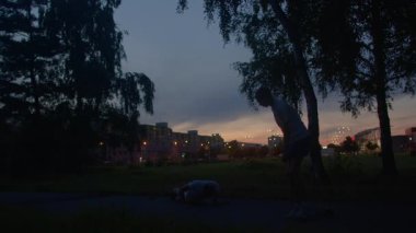Erkek atlet şehir parkının gün batımı yolda yere yığılırken, partner yardım etmek için diz çöküyor, alacakaranlık siluetleri ve ağaç siluetleri çerçeve acil ama sakin açık hava yardım sahnesi, destek ve iyileşmeye odaklanıyor
