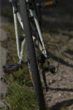 Kapalı bisiklet arka tekerleği ve zinciri kumlu banliyö kıyafetlerini, metaldeki güneş ışığı yansımalarını lastikteki çamur lekelerini, çerçevedeki yıpranmış boyaları, ışığı yakalayan turuncu reflektörleri, sessizliği gösteriyor.