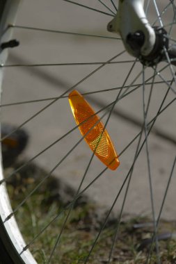 Yakın çekimde bisiklet tekeri yansıtıcısı güneş ışığını tellere yansıtıyor, çimenli kaldırıma çarpıcı turuncu ışık, kentsel hareketlilik ve ürün tasviri için güçlü güvenlik aksanı.