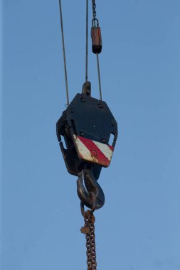 Vinç kanca blok zinciri mavi gökyüzüne asılmış, kırmızı beyaz çizgili yıpranmış çelik, ağır zincir halkaları, kaldırma makara, inşaat alanı ambiyansı, güneş ışığı