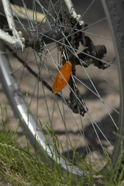 Kapalı bisiklet tekerleği yansıtıcısı ve göbeği, gevrek metal telleri, turuncu mercekli güneşi, ön planda desenli jant ve çim bıçaklarını, özel güvenlik detaylarını gösteriyor.