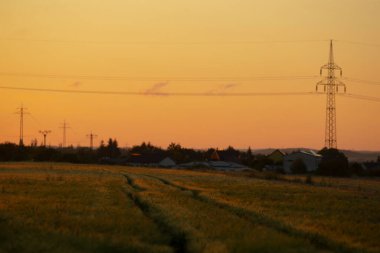 Gün batımı, kuleye doğru izler, uzak köy çatıları üzerinde sıcak turuncu gökyüzü, gerilmiş kablolar ve parlayan ufuklar kamu denetimi ve nostaljik kırsal banliyö sahneleri çağrıştırıyor..