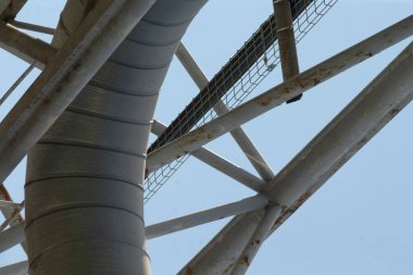 kiriş altındaki kıvrımlı havalandırma kanalı segmentli kaplama ve flanj bağlantılarını gösteriyor, destek parantezleri ve soluk gökyüzüne karşı perçinlenmiş dikişleri, faydacı dokuyu ve mühendislik eklemlerini gösteriyor