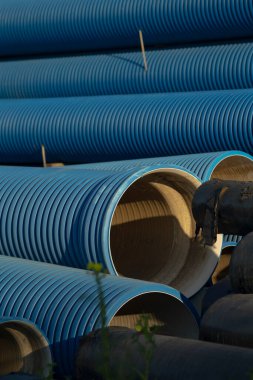 Yığılmış mavi eğimli borular dış depolama alanındaki çakıllara yerleştirilmeyi bekliyor. Katmanları ayıran ahşap uzay araçları, dalgalı yüzeyleri aydınlatan güneş ışığı, siyahla tezat.