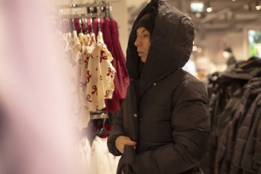 Kadın butikte palto askılığı, kapüşonlu kabarık ceket, asılı elbiseye uzanmak, yumuşak ılık ışık, odaklı ifade, kalabalık perakende koridorları, kumaşlar, mevsimlik kış mevsimi