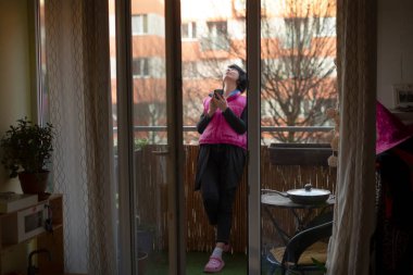 Kadın balkonda fotoğraf çekiyor parlak pembe ceket giyiyor ve spor ayakkabılar giyiyor cam kapıya yaslanıyor saksı bitkileri ve küçük bistro masa dairesi ile çevrili