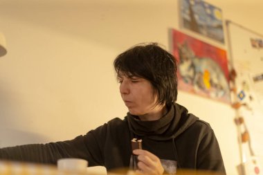 Ev stüdyosunda resim yapan kadın, arka planda tuval, palet ve merdiven üzerinde duran odaklı ifade, duvardaki çerçeveli çalışmalar, yaratıcı süreç ve dokunsal keşif,