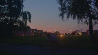 Gün batımı parkı silueti koşucusu huş ağacının altında dinleniyor, şehir ışıkları parlıyor, sakin alacakaranlık silueti, egzersizden sonra yansıtıcı duraklama, hafif lamba ışığı, serinleme esneme, huzurlu kentsel refah titreşimi