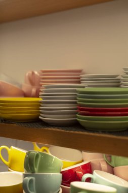 Rafa istiflenmiş renkli bardaklar ve tabaklar, pastel bardaklar ve servis için hazır parlak fincan tabakları ile dolu restoran içi görüntüler, canlı tonlarda kahve ve görsel ortam yaratır.