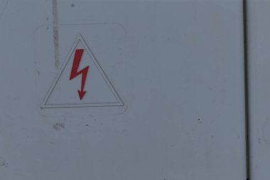 Uyarı işaretli gri elektrik kabini, şehrin dış kısmında bakım noktası, çizik boya ve metal panel, sürgülü ve etiketli kilitli kapı, dikkat üçgeni
