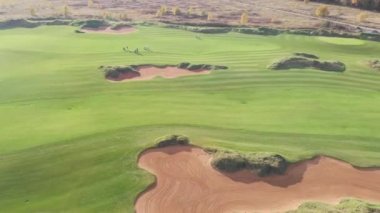 Kırsaldaki bir golf sahasının canlı hava aracı görüntüsü, zengin yeşil bir çim ve kum sığınaklarının olduğu arazi yaz güneşli bir gün, yürüyen ve oynayan bir grup golfçü.