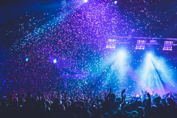 A crowded concert hall with scene stage lights, rock show performance, with people silhouette, colourful confetti explosion fired on dance floor during a concert festival