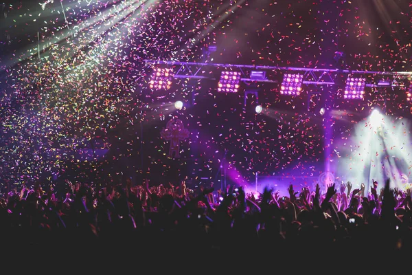 A crowded concert hall with scene stage lights, rock show performance, with people silhouette, colourful confetti explosion fired on dance floor during a concert festival