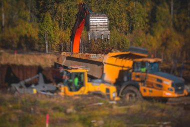 Sarı ağır kazıcı ve çöp kamyonu bahşişçisi kum kazıyor ve yol çalışmaları sırasında çalışıyor, etrafta işçilerin olduğu yeni bir yolun inşaatı sırasında kum boşaltıyor