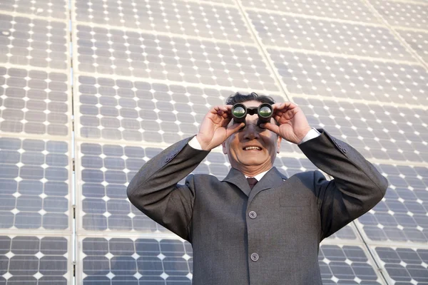 Empresário olhando através de binóculos na frente de painéis solares — Fotografia de Stock