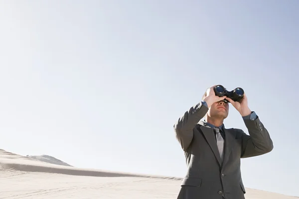 Businessman looking through binoculars in desert — Stock Photo, Image