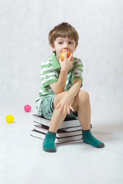 Chłopiec jedzenia jabłko siedzi na stos książek — Zdjęcie stockowe