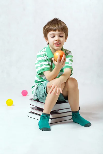Chłopiec jedzenia jabłko siedzi na stos książek — Zdjęcie stockowe