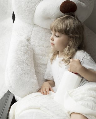 5 yaşındaki bir kız pencereden bakıyor. Arka planda pelüş oyuncak ayı.