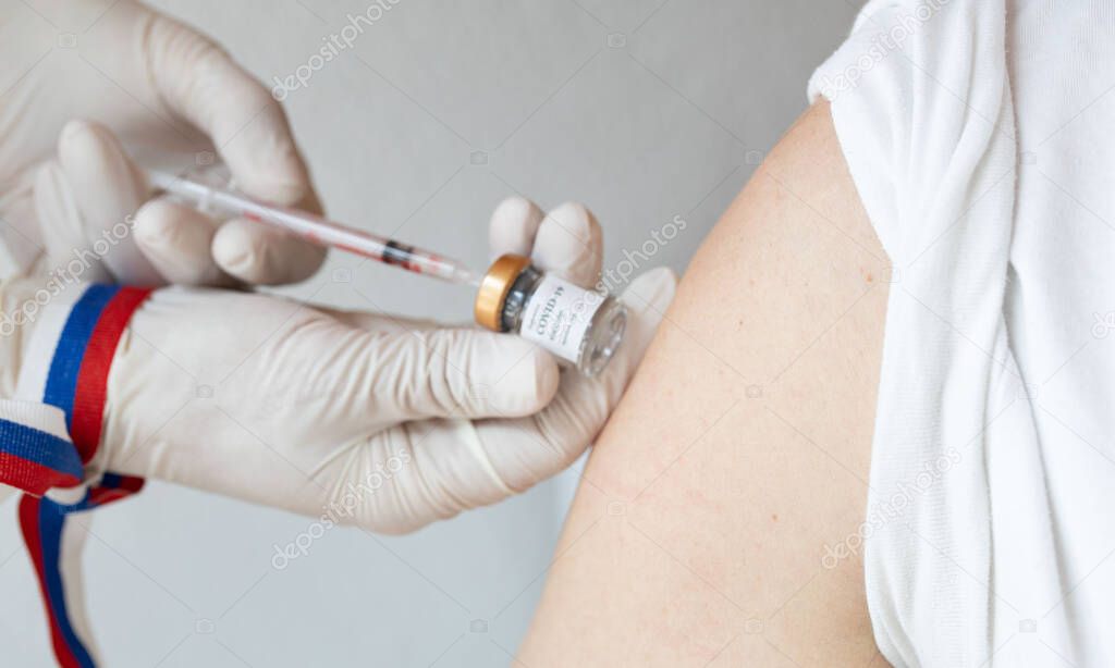 Open shoulder of the man before vaccination. Medical assistant preparing the syringe and Russian flag on her hand in the background.