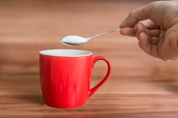 Mão segura colher de chá com açúcar acima da xícara vermelha de chá. Conceito de alimentação não saudável . — Fotografia de Stock