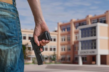 Gun control concept. Young armed man holds pistol in hand in public place near high school. clipart