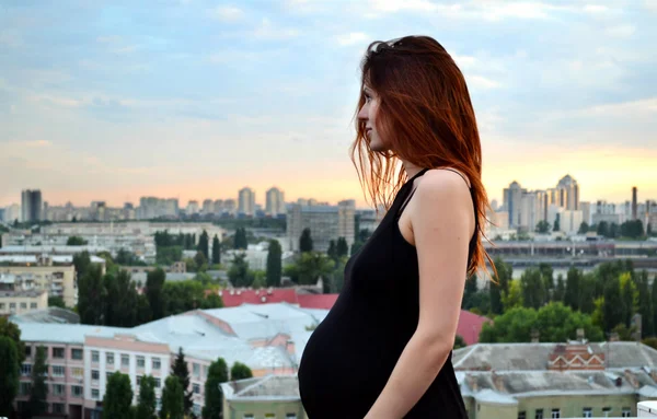 Joven chica pelirroja embarazada hermosa y feliz sobre la vista de la puesta del sol y la ciudad mirando hacia adelante de la maternidad futura y esperando a su bebé en un vestido negro romántico — Foto de Stock