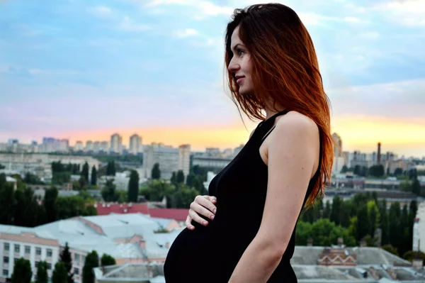 Joven chica pelirroja embarazada hermosa y feliz sobre la vista de la puesta del sol y la ciudad mirando hacia adelante de la maternidad futura y esperando a su bebé en un vestido negro romántico — Foto de Stock