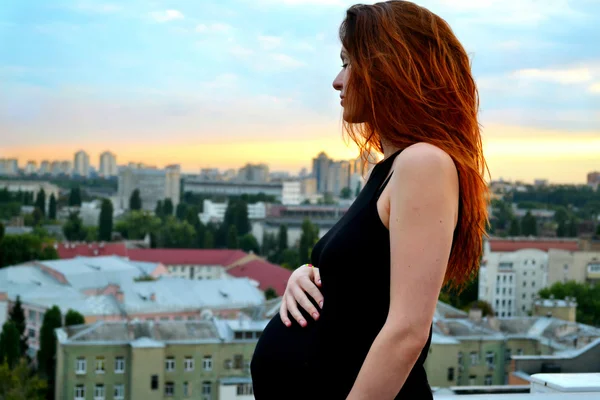Joven chica pelirroja embarazada hermosa y feliz sobre la vista de la puesta del sol y la ciudad mirando hacia adelante de la maternidad futura y esperando a su bebé en un vestido negro romántico — Foto de Stock