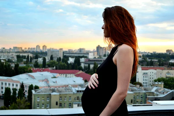 Joven chica pelirroja embarazada hermosa y feliz sobre la vista de la puesta del sol y la ciudad mirando hacia adelante de la maternidad futura y esperando a su bebé en un vestido negro romántico — Foto de Stock