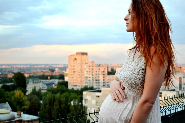 年轻美丽和快乐怀孕红发女孩的日落和市看观提出的未来的母亲和她怀孕在浪漫的白色连衣裙 — 图库照片