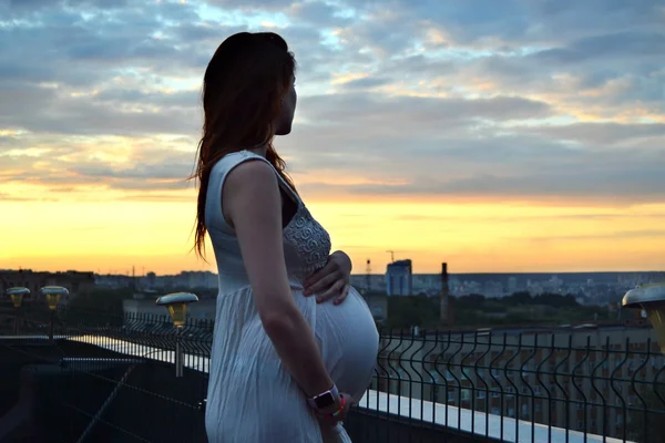 Joven chica pelirroja embarazada hermosa y feliz sobre la vista de la puesta del sol y la ciudad mirando hacia adelante de la maternidad futura y esperando a su bebé en un vestido blanco romántico — Foto de Stock