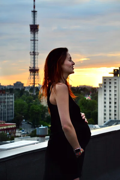 Joven chica pelirroja embarazada hermosa y feliz sobre la vista de la puesta del sol y la ciudad mirando hacia adelante de la maternidad futura y esperando a su bebé en un vestido negro romántico — Foto de Stock