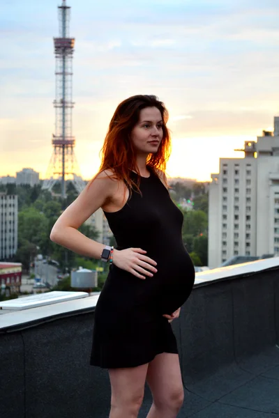 Joven chica pelirroja embarazada hermosa y feliz sobre la vista de la puesta del sol y la ciudad mirando hacia adelante de la maternidad futura y esperando a su bebé en un vestido negro romántico — Foto de Stock