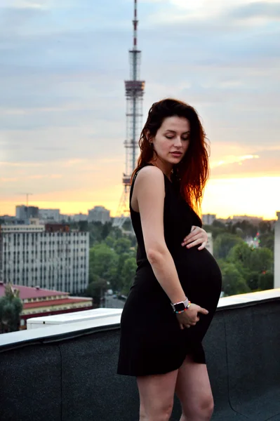 Joven chica pelirroja embarazada hermosa y feliz sobre la vista de la puesta del sol y la ciudad mirando hacia adelante de la maternidad futura y esperando a su bebé en un vestido negro romántico — Foto de Stock