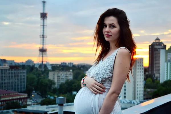 Joven chica pelirroja embarazada hermosa y feliz sobre la vista de la puesta del sol y la ciudad mirando hacia adelante de la maternidad futura y esperando a su bebé en un vestido blanco romántico — Foto de Stock