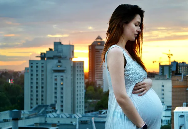 Joven chica pelirroja embarazada hermosa y feliz sobre la vista de la puesta del sol y la ciudad mirando hacia adelante de la maternidad futura y esperando a su bebé en un vestido blanco romántico — Foto de Stock