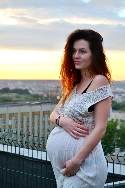 Joven chica pelirroja embarazada hermosa y feliz sobre la vista de la puesta del sol y la ciudad mirando hacia adelante de la maternidad futura y esperando a su bebé en un vestido blanco romántico — Foto de Stock