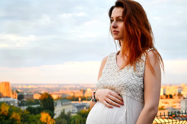 Joven chica pelirroja embarazada hermosa y feliz sobre la vista de la puesta del sol y la ciudad mirando hacia adelante de la maternidad futura y esperando a su bebé en un vestido blanco romántico — Foto de Stock