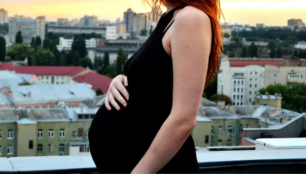 Joven chica pelirroja embarazada hermosa y feliz sobre la vista de la puesta del sol y la ciudad mirando hacia adelante de la maternidad futura y esperando a su bebé en un vestido negro romántico — Foto de Stock