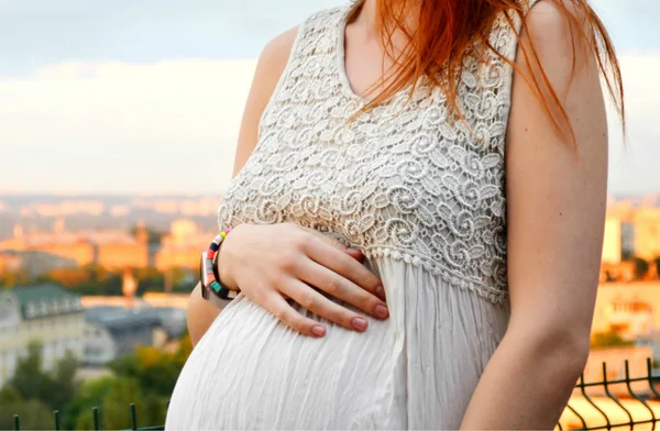 Joven chica pelirroja embarazada hermosa y feliz sobre la vista de la puesta del sol y la ciudad mirando hacia adelante de la maternidad futura y esperando a su bebé en un vestido blanco romántico — Foto de Stock