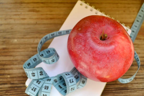 Диетическая концепция с красным яблоком, блокнотом и синей измерительной лентой на деревянном столе — стоковое фото